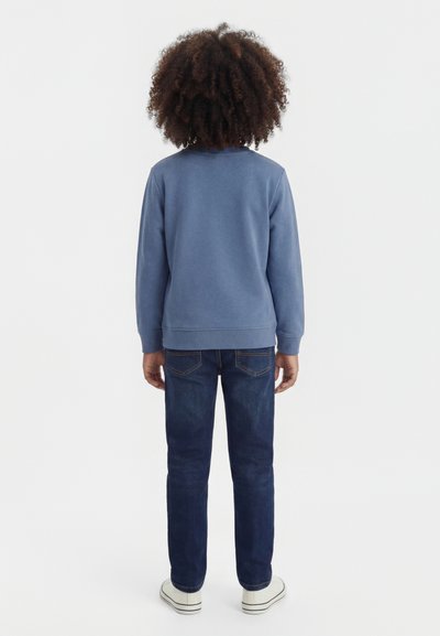 Lokkis juustega laps seisab seljaga, seljas sinine pikkade varrukatega särk, tumedad teksad ja valged tossud ühevärvilise taustaga.