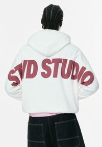 Person med fletninger iført en hvid hættetrøje med stor rød "STUD STUDIO" tekst på ryggen og sorte bukser med synlige lommer.