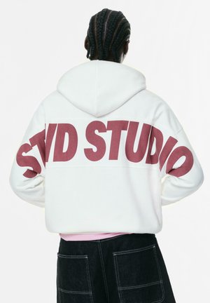 Person with braided hair wearing a white hoodie with large red "STUD STUDIO" text across the back and black pants with visible pockets.