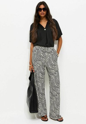 Donna che indossa una blusa nera a maniche corte, pantaloni ampi con stampa a zebra bianco e nero, sandali neri, grandi occhiali da sole, che tiene una borsa tote nera.