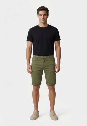 Man staat met het gezicht naar voren, draagt een zwart T-shirt, olijfgroene knielange korte broek en beige sneakers tegen een witte achtergrond.