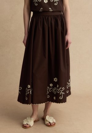 Frau trägt einen braunen Midi-Rock mit weißer Blumenstickerei und gewelltem Saum, kombiniert mit cremefarbenen, gehäkelten Hausschuhen mit Blumenmuster.