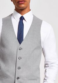 Grauer Weste mit strukturierter Oberfläche, fünf Knöpfen und Schalkarven. Kombiniert mit einem weißen Hemd und einer dunkelblauen Krawatte mit feinem Muster.