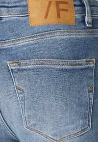 Ljusblå denimjeans med en läderlapp märkt "/F." Har framträdande sömmar på bakfickorna och en texturerad, urblekt tygliknande yta.