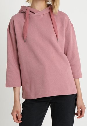 Loose-fit dusty pink hoodie with wide drawstrings, three-quarter sleeves, worn with black pants, midsection and arms visible.