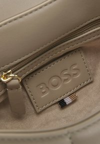 Beige läderväskas inre med en slät textur och guld blixtlås. Har en upphöjd "BOSS"-logotyp och dekorativ stitching i neutrala toner.