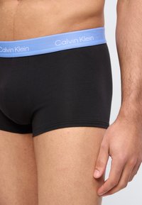 Κοντινό πλάνο στον κορμό και τα πάνω μέρη των ποδιών ενός άνδρα, που φοράει μαύρα ανδρικά εσώρουχα Calvin Klein με ανοιχτόχρωμη γαλάζια ζώνη μέσης.