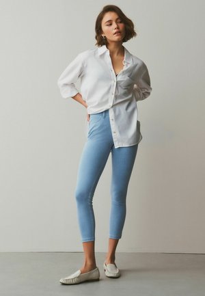 Woman with short brown hair wearing a white button-up shirt, light blue cropped jeans, and white slip-on shoes standing against a plain wall.