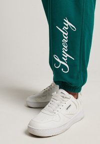 Grüne Jogginghosen haben ein weißes "Superdry"-Logo an der Seite. Kombiniert mit weißen Sneakern, die über perforierte Details und strukturierte Sohlen verfügen.