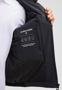 Person hält eine offene schwarze Jacke, die die Innentasche mit Strellson-Logo und Stoffmerkmale-Icons zeigt.