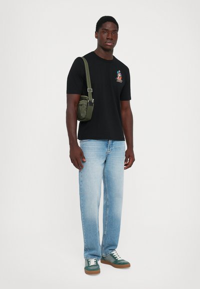 Man wearing black graphic tee, light blue jeans, green sneakers, and carrying small olive green shoulder bag against white background.