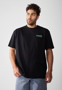 Svart bomull t-skjorte med rund hals, korte ermer, og en grønn "Tropica" logo på venstre bryst. Jevn tekstur, avslappet passform.