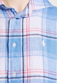 Ljust blå rutigt knäppskjorta tillverkad av tyg med mjuk textur; har röda och svarta ränder, en vit logga och framknappar.