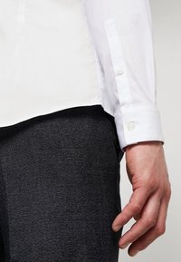 Mano appoggiata accanto a pantaloni neri texture scura, indossando una camicia bianca a maniche lunghe con polsini abbottonati su uno sfondo neutro.