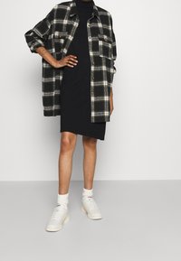 Personne portant une robe noire longueur genou, une chemise surdimensionnée à carreaux noir et blanc, des chaussettes blanches et des baskets blanches, debout devant un fond uni.