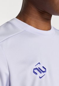 Rövid ujjas, világoslila sportpóló, sima textúrával, kék hímzett logóval a mellkason és kerek nyakkivágással.