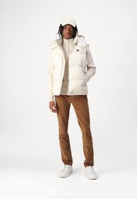 Gilet rembourré de couleur crème avec une capuche, superposé sur un pull beige. Un pantalon en velours côtes marron et des baskets blanches complètent la tenue.