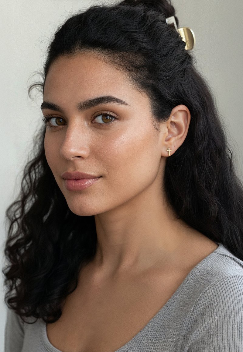 Boucle d'oreille en croix dorée sur un modèle avec de longs cheveux bouclés et foncés, portant un haut côtelé gris clair, avec une peau lisse et un fond neutre.