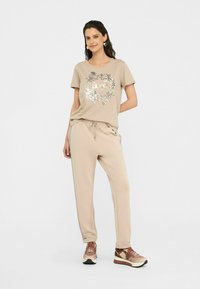 T-shirt beige avec impression florale métallique, associé à un jogging assorti avec taille à cordon. Des chaussures de sport marron et dorées complètent le look.