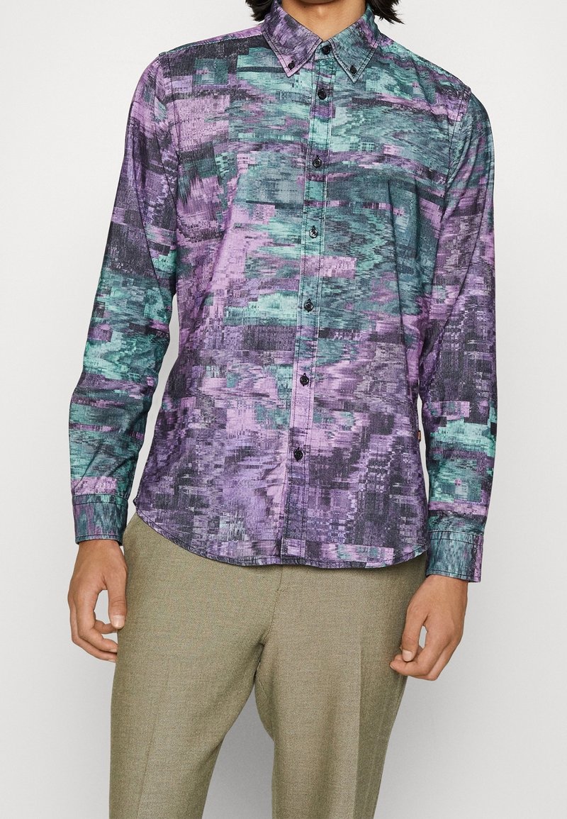 Homme portant une chemise à manches longues boutonnée avec un motif abstrait violet, sarcelle et noir, associée à un pantalon marron clair.