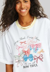 Bílý oversized tričko s barevnými grafickými mašlemi v různých stylech. Tričko má žlutý vroubkovaný výstřih a potištěný text. Měkká látka.