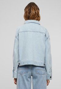 Chaqueta de mezclilla azul claro con un diseño corto, cuello puntiagudo y puños abotonados. Cuenta con costuras visibles y detalle en la sisa de la espalda.