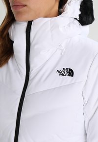 Weiße gepolsterte Jacke mit hohem Kragen, ausgestattet mit einem schwarzen Reißverschluss und Logo. Die Kapuze hat einen Kunstfellbesatz für zusätzliche Details.