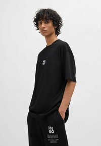 T-shirt oversized noir en tissu doux ; présente un logo blanc "HUGO" sur la poitrine. Le modèle porte un bas noir assorti avec impression de texte.