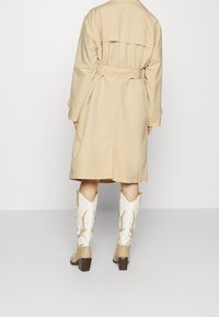 Person wearing a beige trench coat and cream and tan cowboy boots, standing against a plain light gray background, back facing camera.