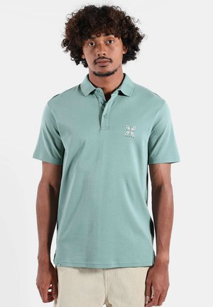 Homme aux cheveux bouclés portant un polo vert clair avec un petit logo blanc brodé, debout devant un fond blanc uni.
