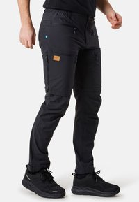 Black outdoor pants with an elastic waistband, multiple zippered pockets, and a brown leather label. Paired with black athletic shoes.