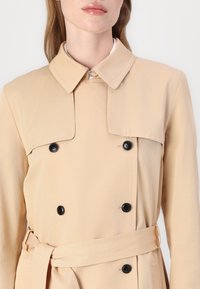 Manteau beige avec un col classique, design croisé à double boutonnage, boutons noirs et ceinture assortie. Texture lisse et coupe ajustée.
