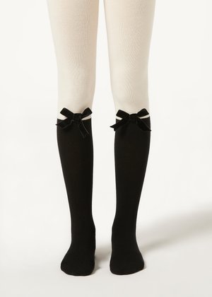 Jambes portant des collants blancs et des chaussettes noires montantes ornées de nœuds en ruban noir en haut, debout sur un fond blanc.