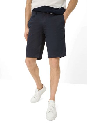 Uomo che indossa pantaloncini blu scuro, scarpe da ginnastica bianche e una maglietta bianca e blu, in piedi con una mano in tasca davanti a uno sfondo bianco.