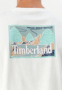 Balta medvilninė marškinėlių su grafiniu dizainu, pavaizduojančiu kalnus ir medžius. Centrinėje dalyje tekstas "Timberland" parašytas ryškiomis raidėmis.