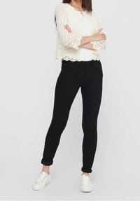 Lace-knit cream cardigan with 3/4 sleeves, paired with black fitted jeans and white sneakers. Features scalloped hem on the cardigan.