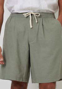 Pantalones cortos de lino verdes con cintura elástica, que presentan un cordón en un color claro y bolsillos laterales. Textura lisa sin patrones visibles.
