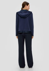 Vrouw die met haar rug naar de camera staat, gekleed in een marineblauwe hoodie en wijde zwarte broeken tegen een eenvoudige lichte achtergrond.