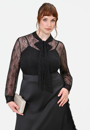 Donna che indossa una blusa nera in pizzo con fiocco e gonna nera che tiene una piccola pochette argento su sfondo bianco.