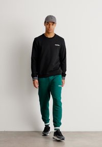 Felpa nera con logo bianco, pantaloni jogger verdi con logo, e cappellino grigio. Look sportivo con accenti bianchi e verdi a contrasto.