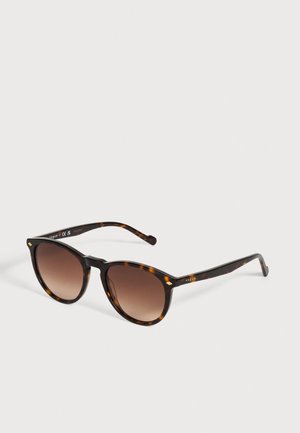 Lunettes de soleil Vogue en écaille de tortue, avec des verres bruns dégradés aux formes arrondies et des détails dorés, sur un fond uni et clair.