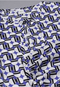 Blusa colorata con un motivo geometrico di cerchi bianchi e neri su uno sfondo blu. Tessuto leggero e liscio con scollatura rilassata.