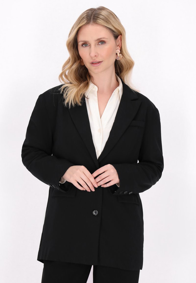 Blazer nero con chiusura a un bottone, revers a notsche e due tasche frontali. Indossato su una camicia bianca, con una texture liscia.