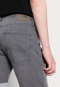 Jeans de mezclilla gris con un diseño de bolsillo trasero, parche con logo en la cinturilla y dobladillo enrollado. Textura suave con detalles de costura sutiles.