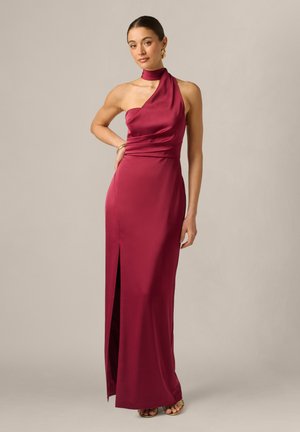 Kvinde iført en ærmeløs, one-shoulder dyb rød satin kjole med højt slids på låret og guld sandaler, stående mod en ensfarvet baggrund.