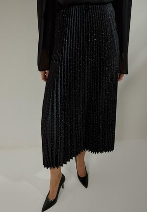 Žena nosí čiernu plisovanú midi sukňu s malými bielymi bodkami, priehľadný čierny dlhý rukáv a čierne špicaté vysoké podpätky, stojí na bielom podlahe.