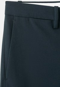 Zbliżenie na ciemnogranatowe, dopasowane spodnie, prezentujące pas biodrowy, szlufkę na pasek oraz szew bocznej kieszeni na jednolitym tle.