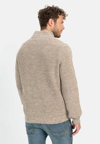 Beige gebreide trui met een hoge kraag en een textuurpatroon. Lange mouwen en een ontspannen pasvorm. Draagt over blauwe jeans, getoond vanuit de achterkant.