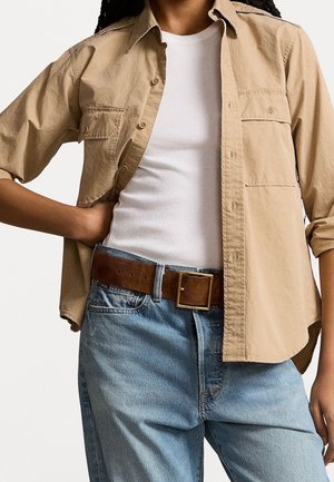Personne portant une chemise beige à boutons sur un débardeur blanc côtelé, un jean bleu et une ceinture en cuir marron avec une boucle carrée en or.