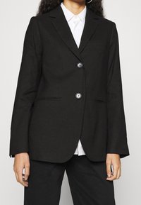 Blazer noir à coupe ajustée, avec deux boutons, col à revers crantés et poches latérales. Le tissu présente une texture lisse et structurée.